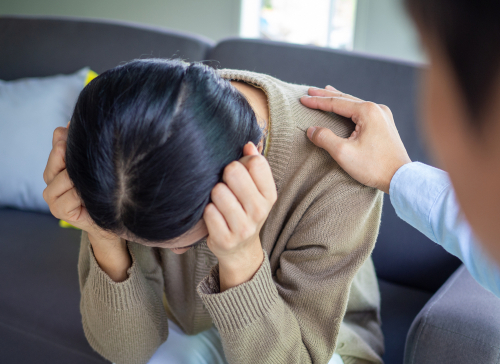 A person sits with their head in their hands, appearing distressed, while another person places a comforting hand on their shoulder.