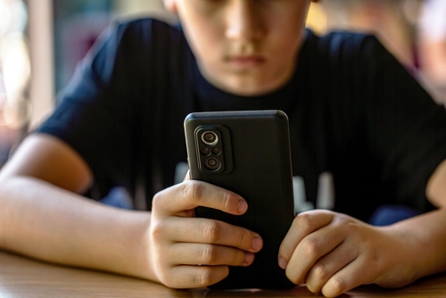 A child sits at a table holding and looking at a smartphone with both hands.