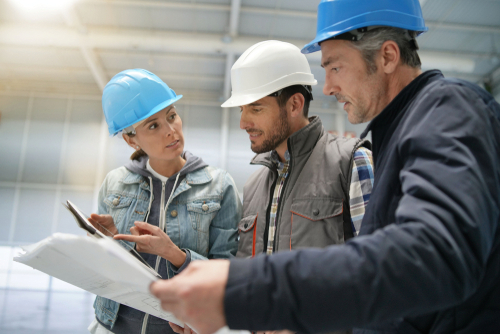 Three construction workers wearing hard hats and casual clothing review blueprints and a clipboard together inside an industrial building.