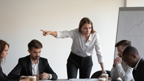 A woman stands and points while addressing four colleagues seated at a table during a meeting, with a chart visible on an easel in the background.