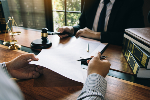 Two people in suits sit at a desk with legal documents, a gavel, scales of justice, and law books, discussing paperwork in an office.