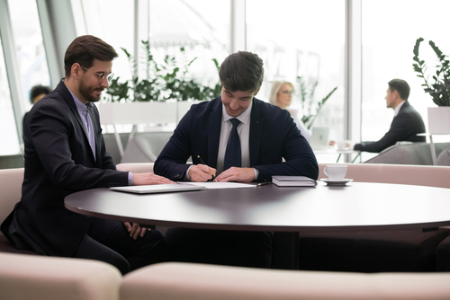 Two men in business suits sitting at a round table, one signing a document while the other points, with colleagues and plants in the background.