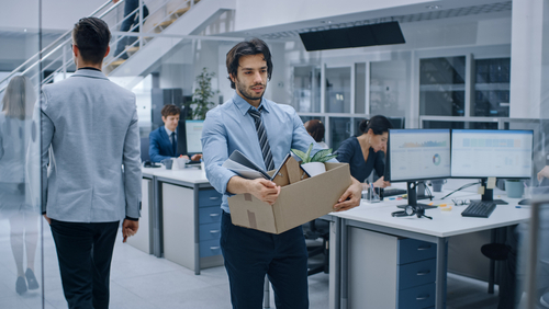 A man in business attire carries a box of personal belongings through an office, suggesting he is leaving his job. Several colleagues work at desks in the background.