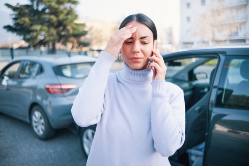 A woman stands outside a car with the door open, holding her forehead and talking on the phone, appearing concerned. Two cars are parked close together in the background.