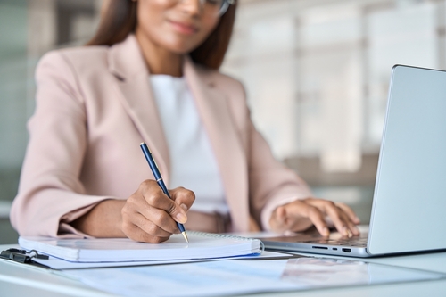 A woman in a light blazer writes in a notebook with one hand while using a laptop with the other, seated at a desk in an office setting.