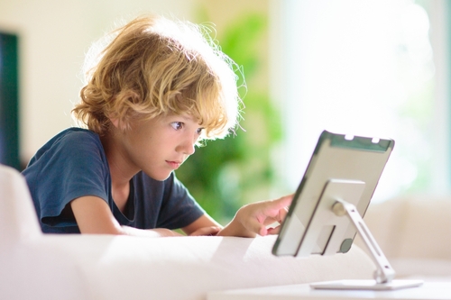 A young boy with blonde hair intently uses a tablet on a stand while sitting on a white sofa in a brightly lit room.
