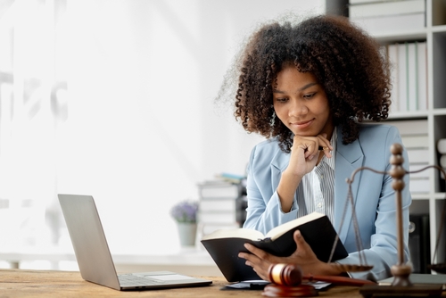 A woman in a blazer reads a book at a desk with a laptop, gavel, and scales of justice in a bright office.
