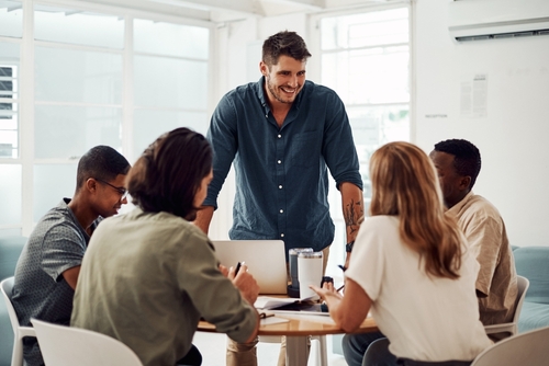 Five people sit around a table in an office, having a discussion. One person stands and smiles while the others listen, with laptops and notebooks on the table.