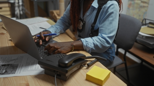 A person sits at a desk working on a laptop, with paperwork, a landline phone, and sticky notes nearby.
