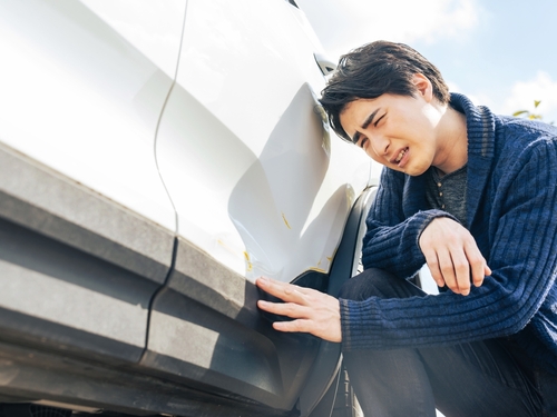 A person kneels beside a white car, examining damage on the rear bumper with a concerned expression.