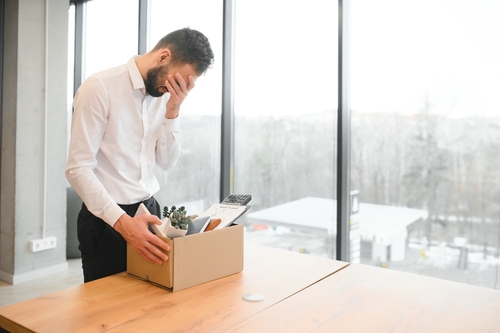 A man in a white shirt stands by a table, covering his face with one hand, looking at a box of personal items, in front of large office windows.