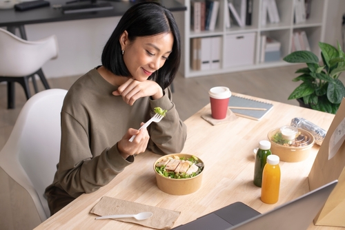 A woman sits at a desk eating a salad from a bowl with a fork, with drinks, a notebook, and a laptop nearby.