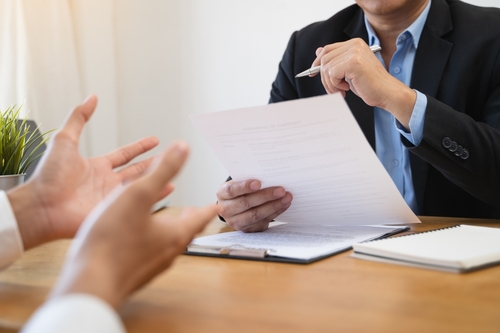 Two people sit across a desk, one holding a document and pen, while the other gestures with their hands during a discussion.
