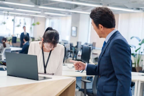 A man in a suit points at a woman standing by a laptop in an office; the woman is bowing her head.
