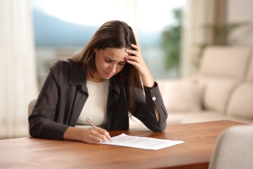 A woman sits at a table, looking concerned while holding her head and writing on a document.
