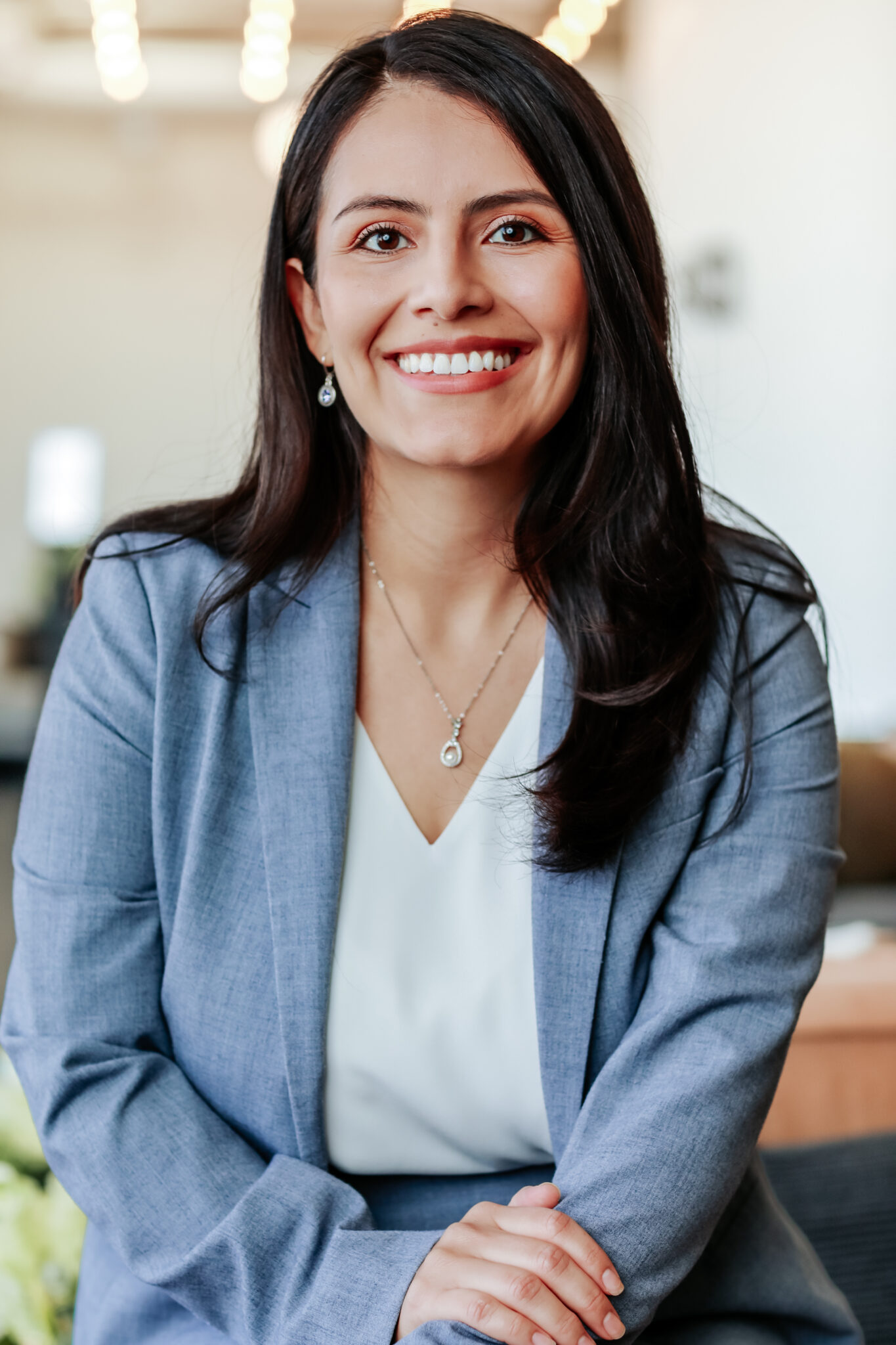 A woman with long dark hair, wearing a light blue suit and white blouse, sits indoors and smiles at the camera.