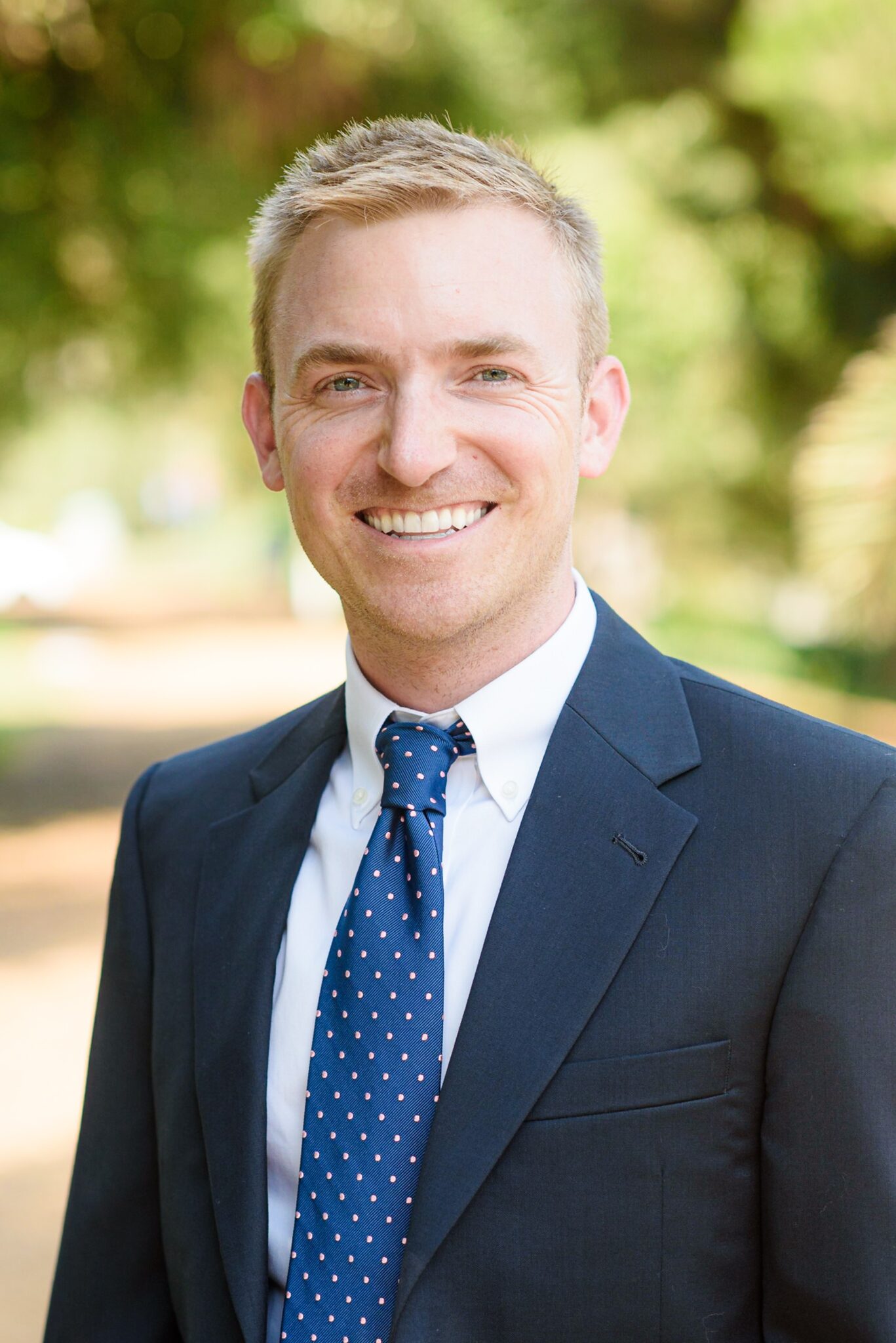 A man in a navy suit and polka dot tie stands outdoors, smiling at the camera with greenery in the blurred background.