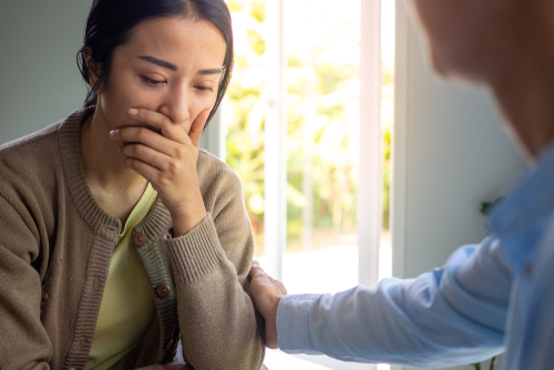 A woman sits with her hand covering her mouth, appearing upset, while another person offers comfort by touching her arm.