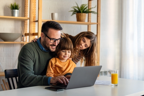 A man, woman, and child sit together at a table looking at a laptop screen with a glass of orange juice nearby.