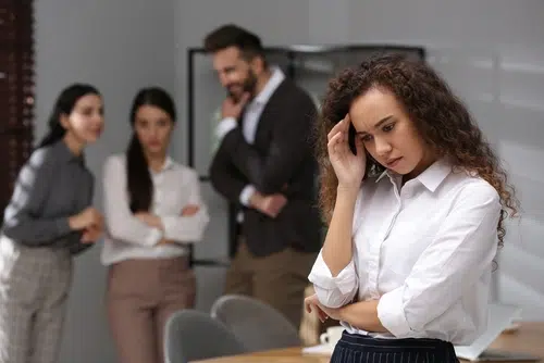 A woman stands apart, looking troubled, while three colleagues in the background appear to be talking and watching her.