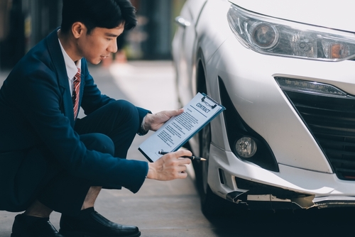 Man in a suit kneels beside a white car, holding a clipboard and pen, inspecting the front bumper and taking notes.