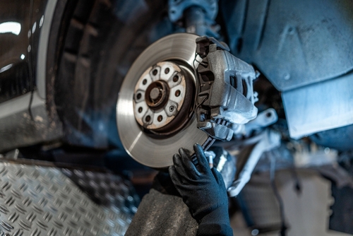 Mechanic wearing black gloves inspecting or installing a car disc brake on a raised vehicle in an auto repair shop.