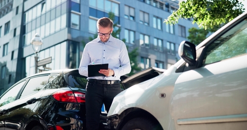 A man stands between two cars involved in a collision, taking notes on a clipboard, with modern buildings and trees in the background.
