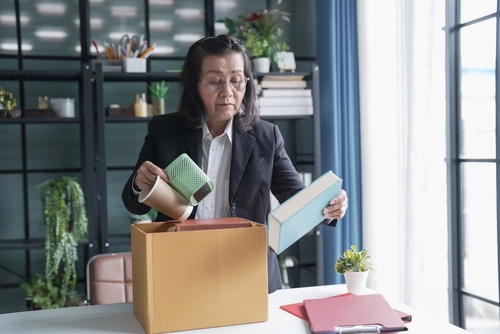 A woman in business attire packs a mug and a book into a cardboard box on a desk, with office shelves and a window in the background.