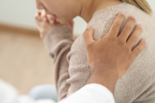 A person places a comforting hand on another person's shoulder, who is sitting and appears to be in distress.