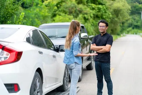A woman stands beside a white car, talking to a man on a road with trees in the background; a black vehicle is parked behind them.
